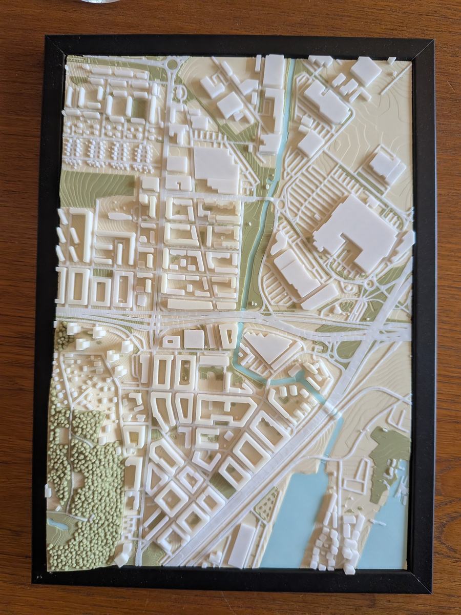 Foto: ein gedrucktes Stadtmodell in einem schwarzen Bilderrahmen, von oben – weiße Gebäude auf cremefarbenem Grund, ein hellblauer Fluss und See, grüne Parks und ein Wald aus einzelnen Baumkronen.